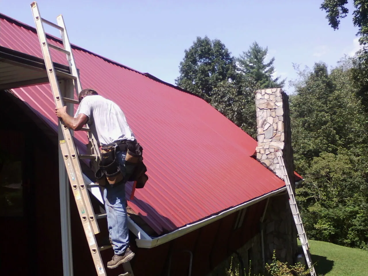 Roofer on a ladder beside a red metal roof and stone chimney — Soffit & Fascia Repair by Roofing Brothers in Boardman